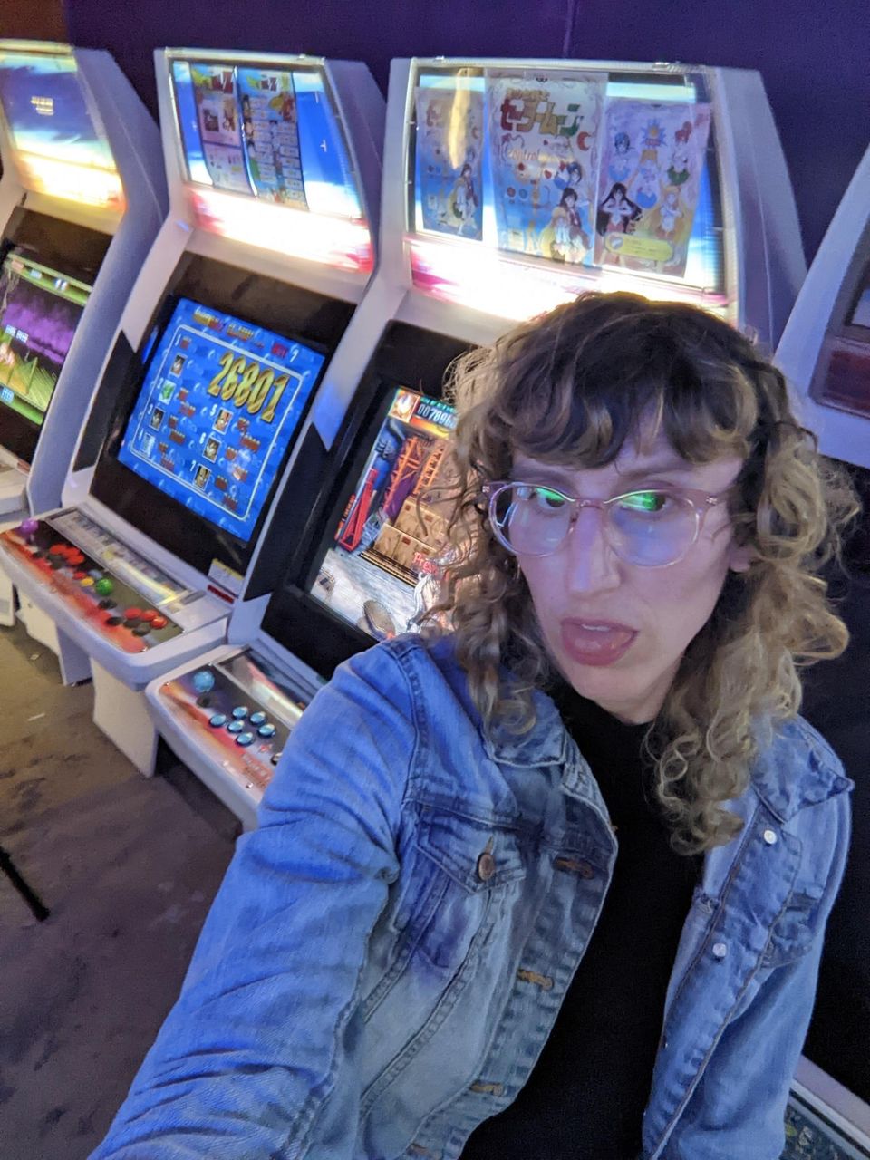 daphnis sitting in front of a row of arcade machines, sticking their tongue out 