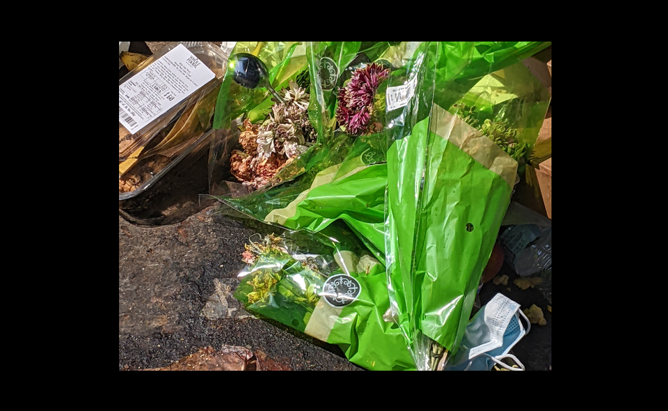 a photo of dried flowers wrapped in plastic at the bottom of a dumpster. there's a mask and a whole foods container